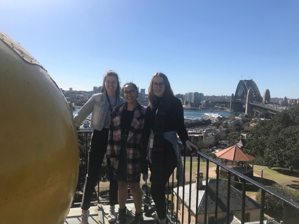 The museum interns stand beside a large yellow ball on a high tower. The Sydney Harbour Bridge is in the background. 